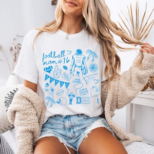 May include: A white short-sleeved t-shirt featuring a blue graphic print with the text football mom #16. The design includes various football-themed illustrations such as a player, a helmet, a whistle, a pennant, a foam finger, and a stadium.