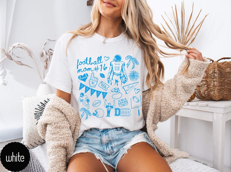 May include: A white short-sleeved t-shirt featuring a blue graphic print with the text football mom #16. The design includes various football-themed illustrations such as a player, a helmet, a whistle, a pennant, a foam finger, and a stadium.