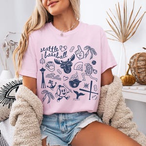 May include: Pink t-shirt with the words "Seattle Baseball" and various navy blue illustrations, including a moose, baseball, and the Space Needle. The shirt is worn with denim shorts and a cream-colored cardigan.