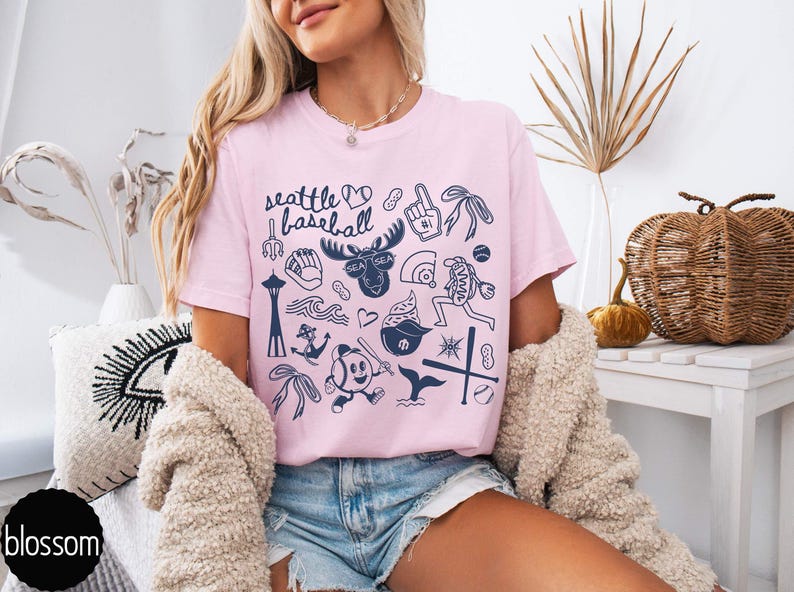 May include: Pink t-shirt with the words "Seattle Baseball" and various navy blue illustrations, including a moose, baseball, and the Space Needle. The shirt is worn with denim shorts and a cream-colored cardigan.