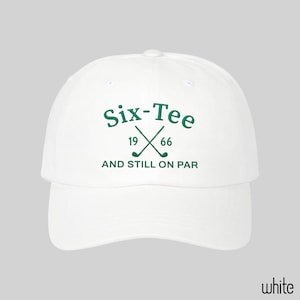 May include: A white baseball cap featuring dark green embroidered text that reads Six-Tee, 19 66, and AND STILL ON PAR, with a graphic of two crossed golf clubs positioned between the numbers 19 and 66 on the front center of the crown.