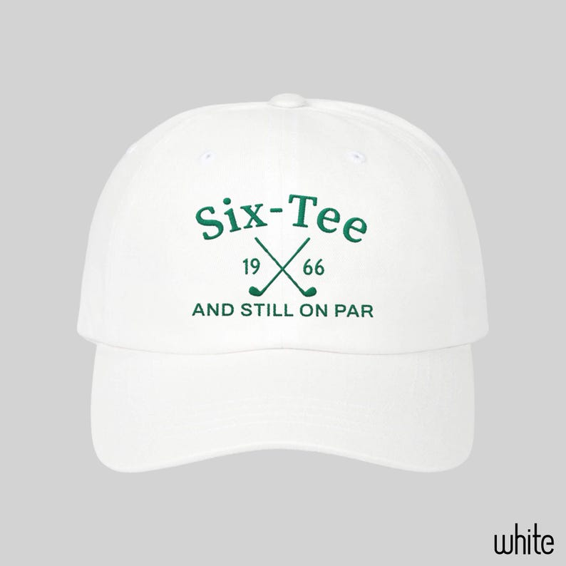 May include: A white baseball cap featuring dark green embroidered text that reads Six-Tee, 19 66, and AND STILL ON PAR, with a graphic of two crossed golf clubs positioned between the numbers 19 and 66 on the front center of the crown.