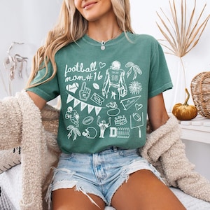 May include: A light green t-shirt featuring white graphic illustrations of football-themed icons, including a player, a helmet, a pennant, a whistle, and the text "football mom #16". The shirt has a crew neckline and short sleeves.