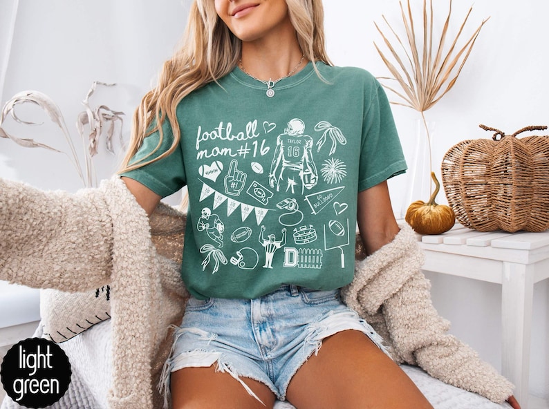 May include: A light green t-shirt featuring white graphic illustrations of football-themed icons, including a player, a helmet, a pennant, a whistle, and the text "football mom #16". The shirt has a crew neckline and short sleeves.