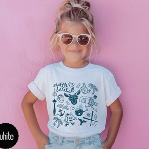 May include: White t-shirt with rolled-up sleeves featuring a teal graphic design of Seattle-themed illustrations, including the Space Needle, a moose, and baseball-related images. The text "Seattle Baseball" is also included.