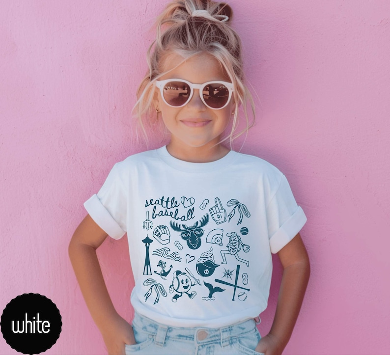 May include: White t-shirt with rolled-up sleeves featuring a teal graphic design of Seattle-themed illustrations, including the Space Needle, a moose, and baseball-related images. The text "Seattle Baseball" is also included.