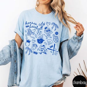 May include: Pale blue t-shirt with a blue graphic design. The design features the text "Kansas City Baseball" and baseball-themed illustrations, including baseballs and crowns. The shirt is made of a soft material. The design is in blue.