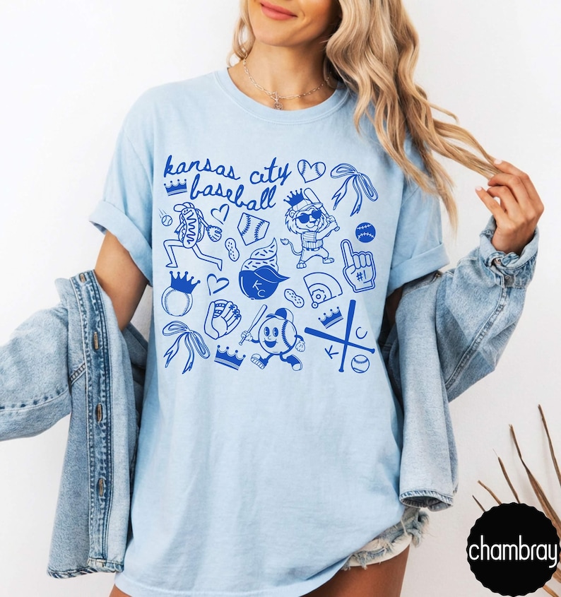 May include: Pale blue t-shirt with a blue graphic design. The design features the text "Kansas City Baseball" and baseball-themed illustrations, including baseballs and crowns. The shirt is made of a soft material. The design is in blue.