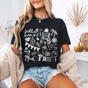 May include: A black short-sleeve t-shirt featuring white line art graphics related to American football, including the text "football mom #53", a player jersey with the number 53, a whistle, a pennant, a football, and various game-day icons.