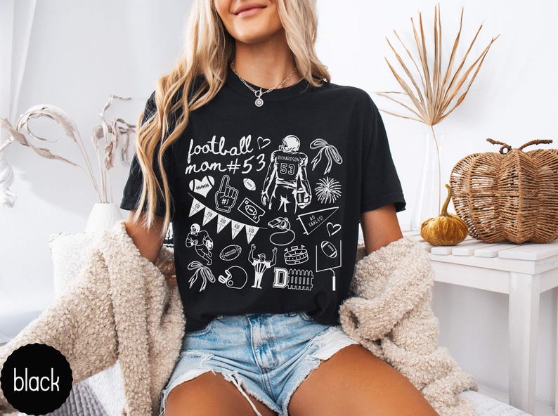 May include: A black short-sleeve t-shirt featuring white line art graphics related to American football, including the text "football mom #53", a player jersey with the number 53, a whistle, a pennant, a football, and various game-day icons.