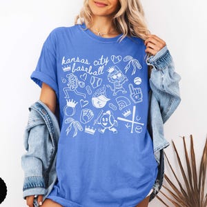 May include: A blue t-shirt featuring the text "Kansas City Baseball" and white baseball-themed illustrations. The shirt has a relaxed fit and is a vibrant shade of blue.