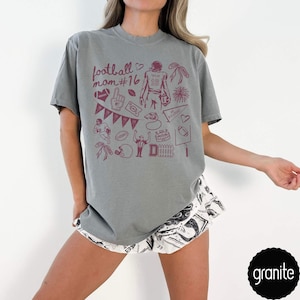 May include: A grey short-sleeved t-shirt featuring a maroon graphic print with the text football mom #16. The design includes illustrations of a football player, a helmet, a pennant, a whistle, a stadium, and various football-themed icons.