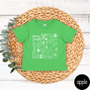 May include: A green toddler t-shirt with a white graphic design featuring the words "Green Bay Football" and various football-related icons. The shirt is displayed on a woven, circular placemat.