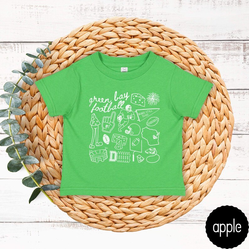 May include: A green toddler t-shirt with a white graphic design featuring the words "Green Bay Football" and various football-related icons. The shirt is displayed on a woven, circular placemat.