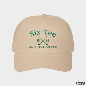 May include: A stone-colored baseball cap featuring dark green embroidery on the front. The text reads Six-Tee 19 66 AND STILL ON PAR, accompanied by an icon of two crossed golf clubs. The cap has a curved brim and a structured crown design.