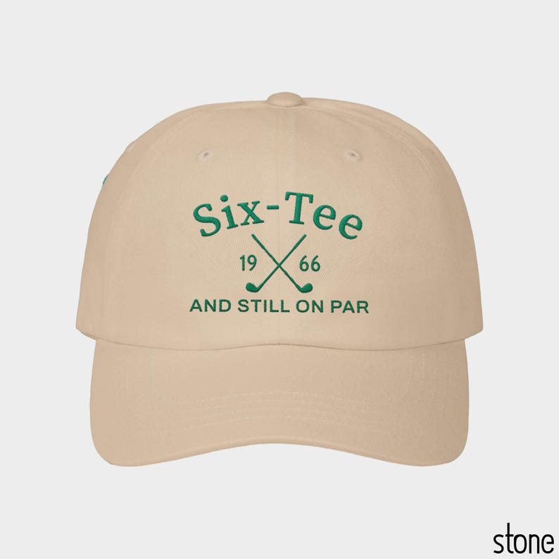 May include: A stone-colored baseball cap featuring dark green embroidery on the front. The text reads Six-Tee 19 66 AND STILL ON PAR, accompanied by an icon of two crossed golf clubs. The cap has a curved brim and a structured crown design.