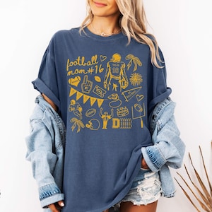May include: A denim blue short-sleeve t-shirt featuring a yellow graphic print with the text football mom #16. The design includes various football-themed illustrations such as a player, a helmet, a foam finger, a pennant, and a goal post.