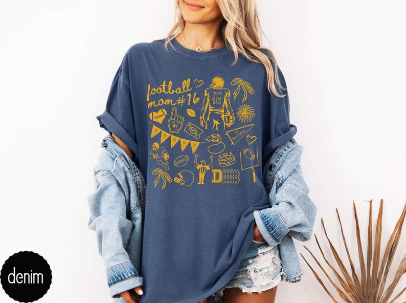 May include: A denim blue short-sleeve t-shirt featuring a yellow graphic print with the text football mom #16. The design includes various football-themed illustrations such as a player, a helmet, a foam finger, a pennant, and a goal post.