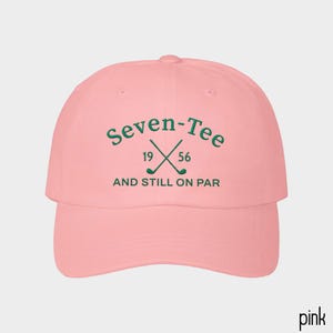 May include: A light pink baseball cap featuring dark green embroidered text that reads Seven-Tee 19 56 AND STILL ON PAR, with a graphic of two crossed golf clubs positioned between the numbers 19 and 56 on the front center of the crown.