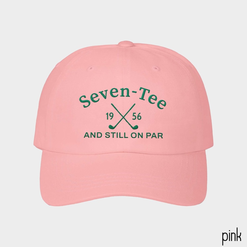 May include: A light pink baseball cap featuring dark green embroidered text that reads Seven-Tee 19 56 AND STILL ON PAR, with a graphic of two crossed golf clubs positioned between the numbers 19 and 56 on the front center of the crown.