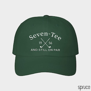 May include: A dark green baseball cap featuring white embroidered text that reads Seven-Tee 19 56 AND STILL ON PAR, accompanied by a graphic of two crossed golf clubs centered between the numbers 19 and 56 on the front panel of the hat.