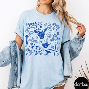 May include: Light blue t-shirt with a blue graphic design featuring the text "Seattle Baseball" and various baseball-themed illustrations. The design includes a baseball, a moose, and other Seattle-related icons.