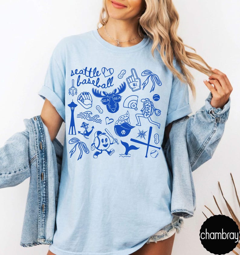 May include: Light blue t-shirt with a blue graphic design featuring the text "Seattle Baseball" and various baseball-themed illustrations. The design includes a baseball, a moose, and other Seattle-related icons.