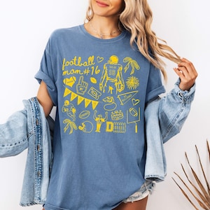 May include: A blue jean colored t-shirt featuring a yellow graphic print with the text football mom #16. The design includes various football-themed illustrations such as a player, a helmet, a pennant, a foam finger, and a stadium.