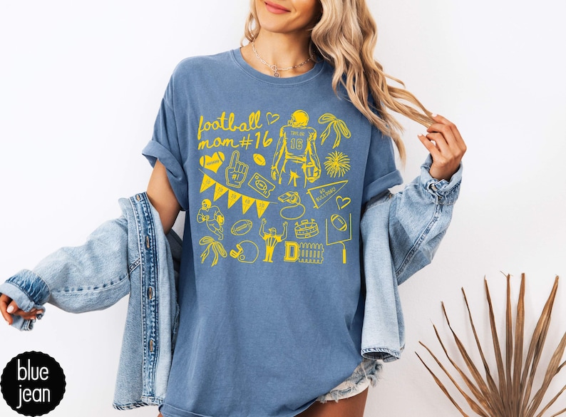 May include: A blue jean colored t-shirt featuring a yellow graphic print with the text football mom #16. The design includes various football-themed illustrations such as a player, a helmet, a pennant, a foam finger, and a stadium.