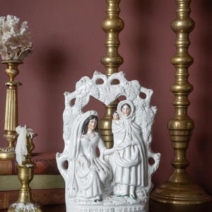 Op de afbeelding: Een wit keramieken beeldje met twee vrouwen en een kind, met een decoratieve druivenboog. De figuren dragen lange witte gewaden. Twee messing kandelaars met witte kaarsen op de achtergrond.