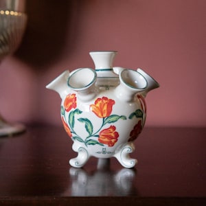 May include: A white ceramic tulipiere vase with a rounded body, multiple spouts, and three scrolled feet. The surface is decorated with hand-painted orange and yellow tulips featuring green stems and leaves, accented by thin green trim lines.