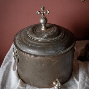 May include: Antique metal water jug with a cross-shaped finial on the lid. The jug has a spigot and decorative handles. The metal has a dark, aged patina. The jug is sitting on a white cloth.