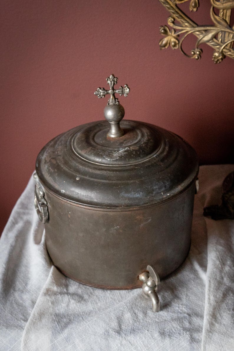 May include: Antique metal water jug with a cross-shaped finial on the lid. The jug has a spigot and decorative handles. The metal has a dark, aged patina. The jug is sitting on a white cloth.