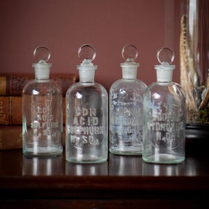 Peut inclure: Quatre flacons d'apothicaire en verre transparent avec bouchons en verre. Chaque flacon porte une étiquette avec du texte. Les flacons sont disposés sur une surface en bois foncé. L'arrière-plan comprend un mur bordeaux et un dôme en verre.