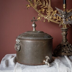 May include: A vintage metal water container, featuring a cross-shaped lid finial and a spigot. It rests on a white cloth, with a decorative gold candelabra in the background. The image evokes a sense of history and antique design.