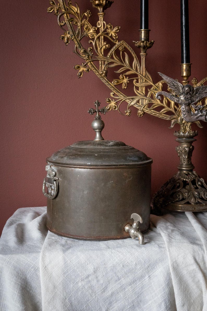 May include: A vintage metal water container, featuring a cross-shaped lid finial and a spigot. It rests on a white cloth, with a decorative gold candelabra in the background. The image evokes a sense of history and antique design.