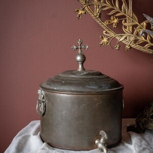 May include: A vintage metal container with a cross-shaped finial on the lid. The container has a tap and is set on a white cloth. A decorative gold wall sconce is in the background.