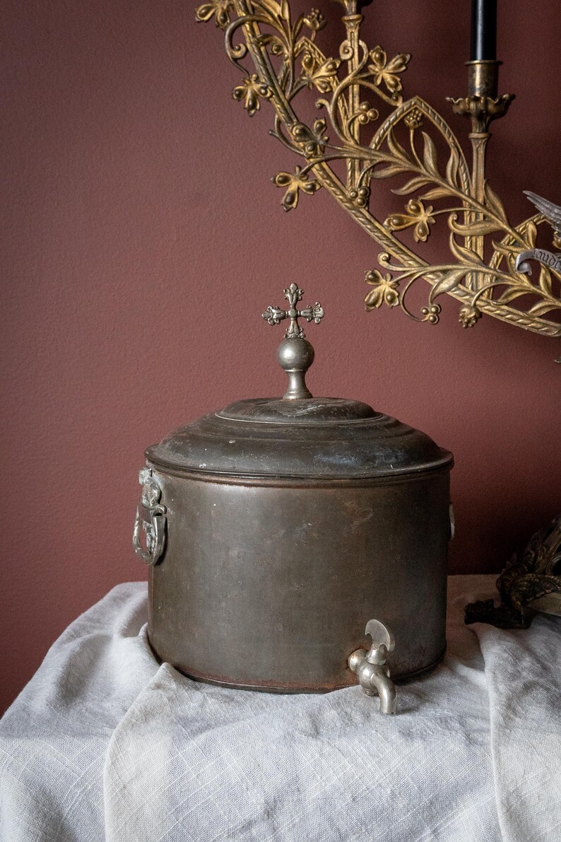 May include: A vintage metal container with a cross-shaped finial on the lid. The container has a tap and is set on a white cloth. A decorative gold wall sconce is in the background.