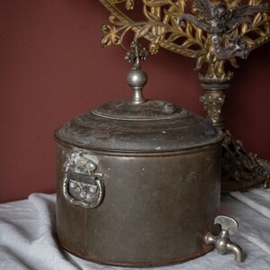 May include: An antique metal water container with a tap, handle, and decorative lid. The cylindrical container has a tarnished finish. A decorative metal candelabra is in the background.