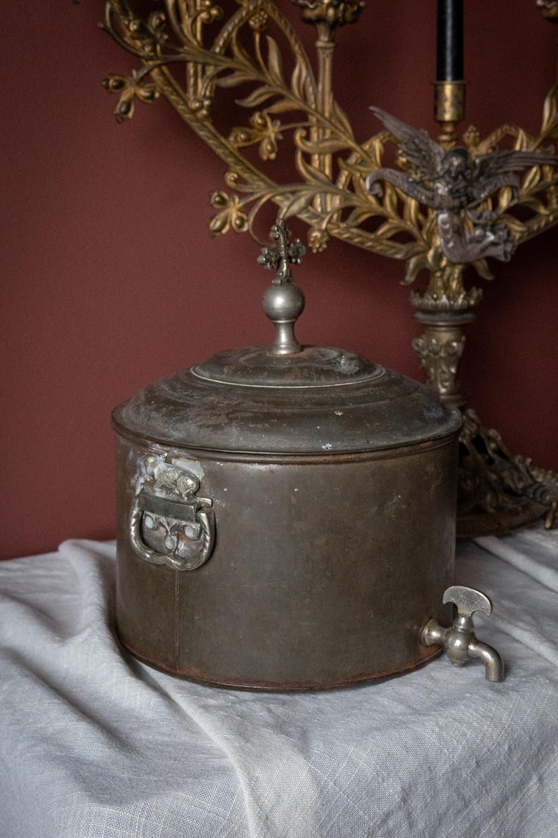 May include: An antique metal water container with a tap, handle, and decorative lid. The cylindrical container has a tarnished finish. A decorative metal candelabra is in the background.