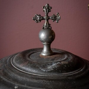 May include: Close-up of a dark metal object with a decorative cross on top. The cross is ornate and sits atop a spherical base. The object has a circular, ridged design and a distressed finish, suggesting age.