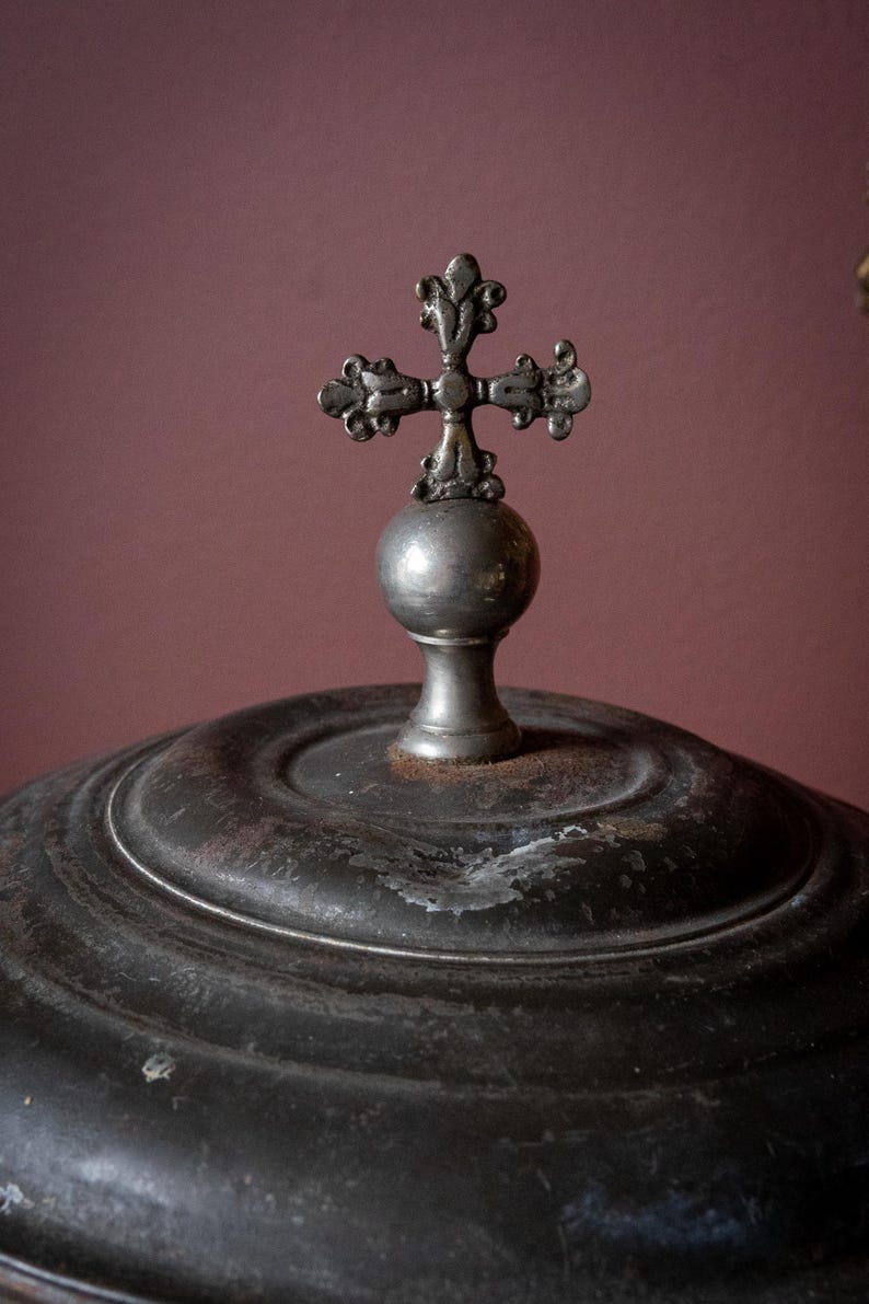 May include: Close-up of a dark metal object with a decorative cross on top. The cross is ornate and sits atop a spherical base. The object has a circular, ridged design and a distressed finish, suggesting age.