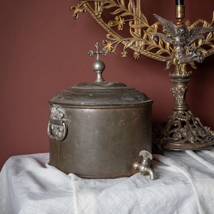 May include: An antique metal container with a lid and a small tap. The container has a cross-shaped finial on top and a handle on the side. A decorative candle holder is in the background.