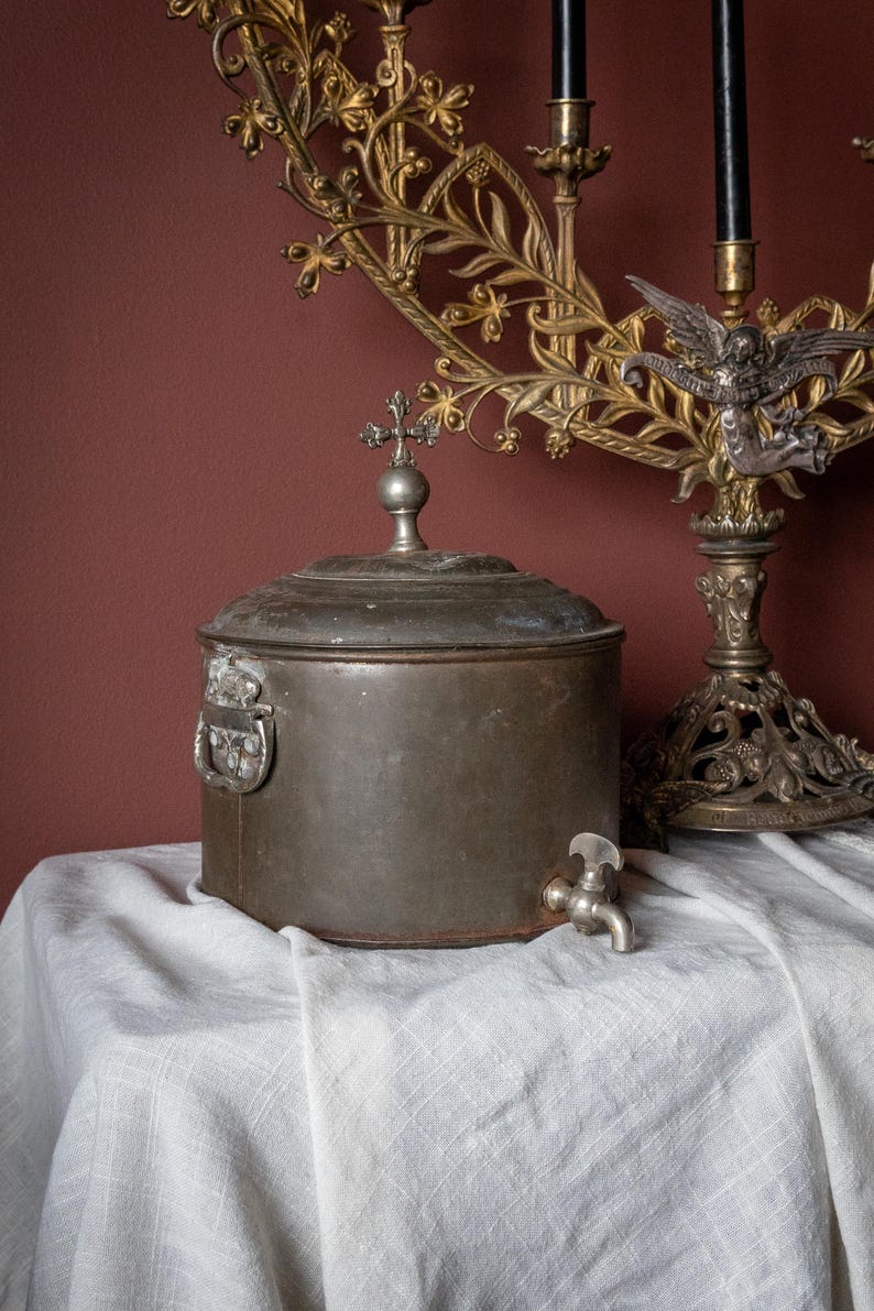 May include: An antique metal container with a lid and a small tap. The container has a cross-shaped finial on top and a handle on the side. A decorative candle holder is in the background.
