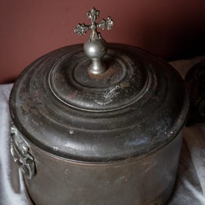 May include: Antique metal container with a lid and a spigot. The container is dark grey with a cross-shaped finial on top. The container has a round shape and is sitting on a white cloth.
