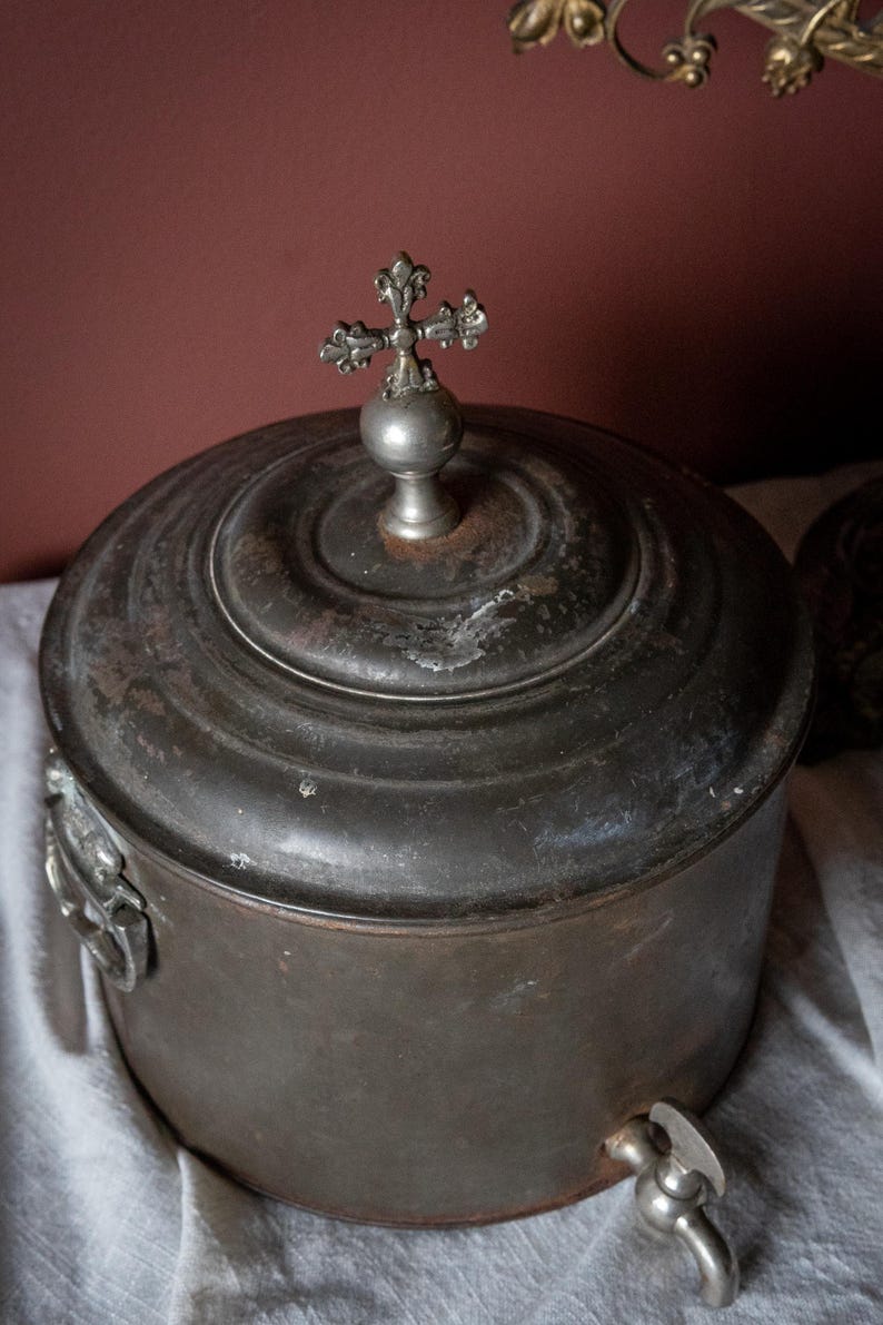 May include: Antique metal container with a lid and a spigot. The container is dark grey with a cross-shaped finial on top. The container has a round shape and is sitting on a white cloth.