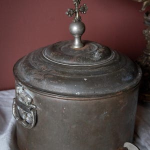 May include: An antique metal container with a lid and a decorative cross finial. The container has a spigot and a handle. The metal has a dark, aged patina, suggesting it is an old piece. Likely used for holding liquids.