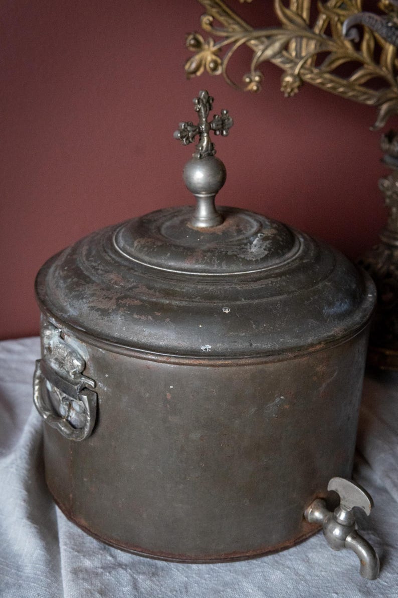 May include: An antique metal container with a lid and a decorative cross finial. The container has a spigot and a handle. The metal has a dark, aged patina, suggesting it is an old piece. Likely used for holding liquids.