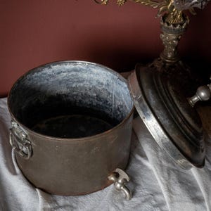 May include: An antique metal container with a lid, tap, and handles. The container is made of dark, aged metal with a silver interior. The lid is partially open, showing the inside. The tap is near the base.