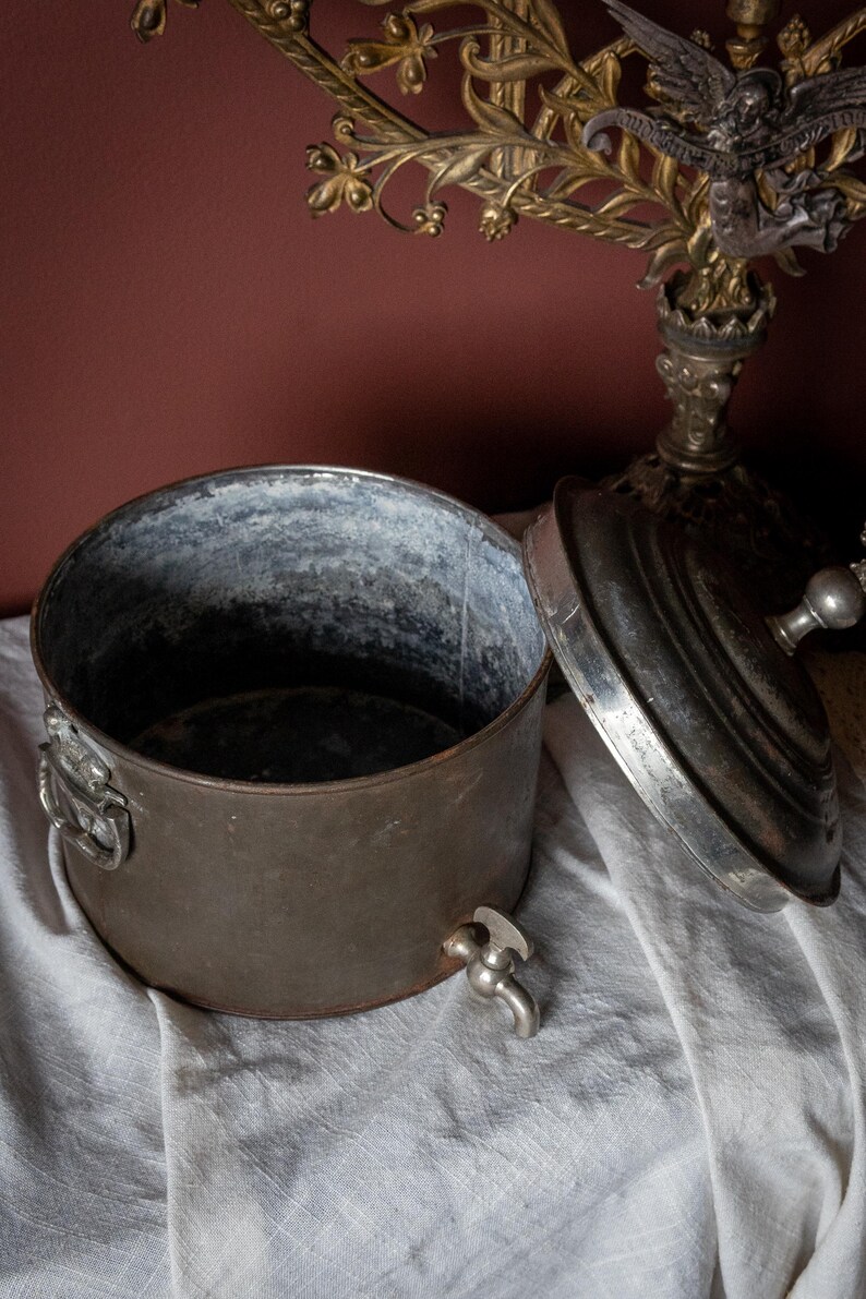 May include: An antique metal container with a lid, tap, and handles. The container is made of dark, aged metal with a silver interior. The lid is partially open, showing the inside. The tap is near the base.